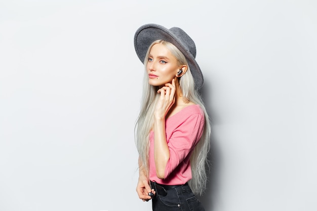 Photo studio portrait of young pretty blonde girl with wireless earbuds in ear wearing grey hat and pink shirt on white background