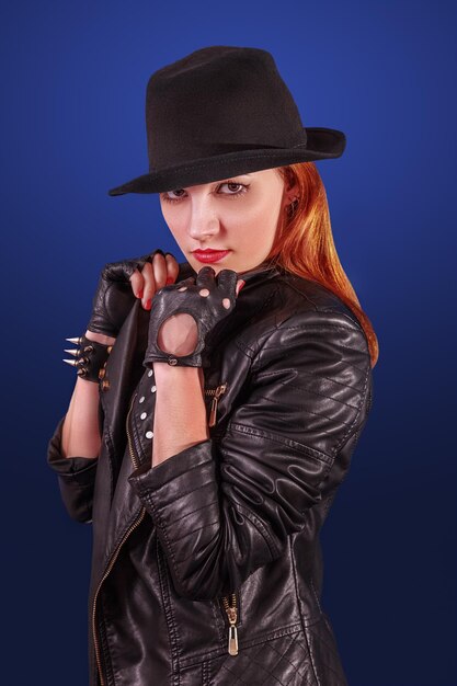 Studio portrait of a young pretty woman in a black hat and\
leather jacket on a blue background