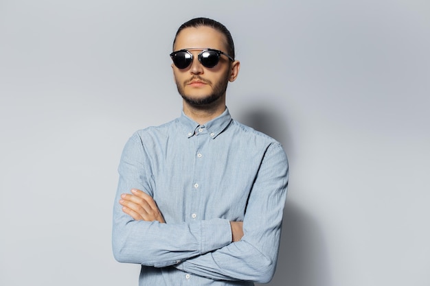 Studio portrait of young serious man with crossed arms wearing sunglasses and blue shirt on white background