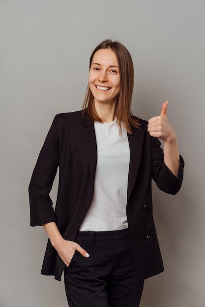 Foto ritratto in studio di una giovane donna sorridente e sicura di sé che tiene un grosso pollice in su su sfondo grigio