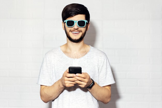 Studio portrait of young smiling guy with smartphone in hands, wearing cyan shades on white