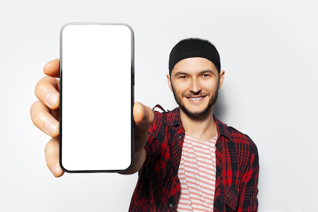 Studio portrait of young smiling man holding big smartphone with blank on screen in hand showing close to camera a device with mockup on white background Wearing red plaid tshirt and striped shirt