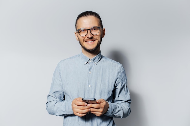 Studio portrait of young smiling man holding smartphone in hands on light grey background Wearing eyeglasses and tshirt