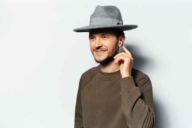 Студийный портрет молодого улыбающегося мужчины, слушающего музыку через беспроводные наушники на белом фоне в свитере и серой шляпе