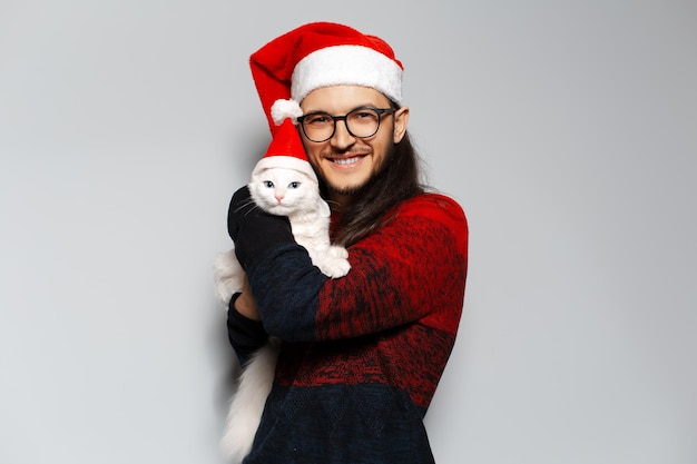 Studio portrait of young smiling man in red sweater hugs white cat wearing Santa hat on white background Christmas concept