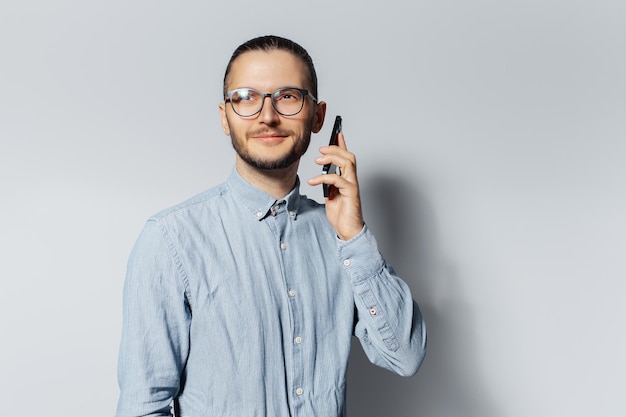 Ritratto in studio di giovane uomo sorridente che parla su smartphone con occhiali da vista e camicia blu su sfondo bianco