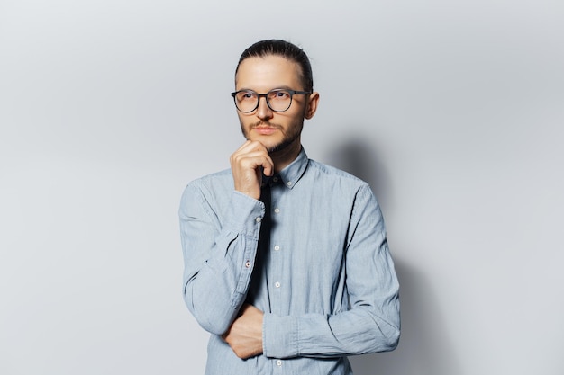 Ritratto in studio di giovane uomo premuroso in camicia blu che indossa occhiali da vista toccando il mento su sfondo bianco