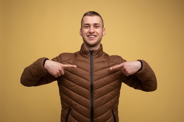 Studio portret. Jonge 20s man in een bruin jasje, op een gele achtergrond, glimlacht en toont zichzelf met zijn vingers