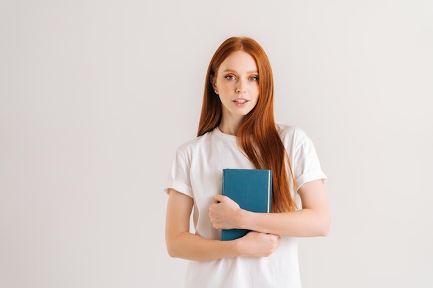 Studio portret van aantrekkelijke jonge student vrouw in handen papieren boek te houden en kijken naar camera staande op een witte geïsoleerde achtergrond Gelukkig roodharige dame poseren met educatief materiaal