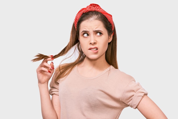 Studio portret van doordachte mooie jonge vrouw draagt beige tshirt en rode hoofdband spelen met een stuk haar ziet er onzeker opzij poses tegen witte achtergrond mensen emoties concept