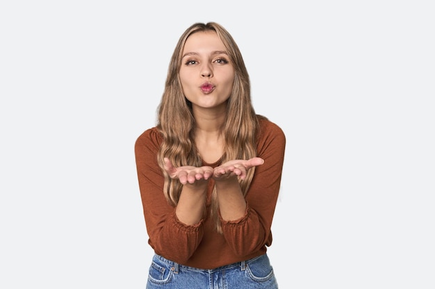 Studio portret van een blonde blanke vrouw die lippen vouwt en handpalmen vasthoudt om een luchtkus te sturen