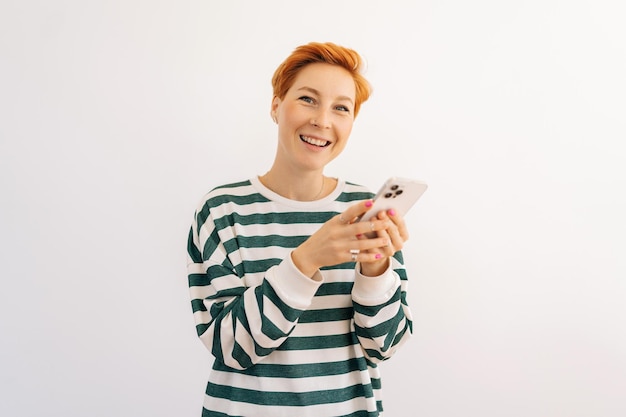 Foto studio portret van een gelukkige aantrekkelijke jonge vrouw in casual kleding die staat met een mobiele telefoon en een sms-bericht schrijft en glimlacht en naar de camera kijkt