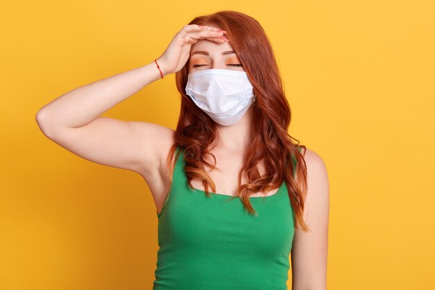 Studio portret van een jong roodharige meisje met gesloten ogen, met hoofdpijn, hand op het voorhoofd houden, medische griep masker en groene t-shirt op gele achtergrond dragen.