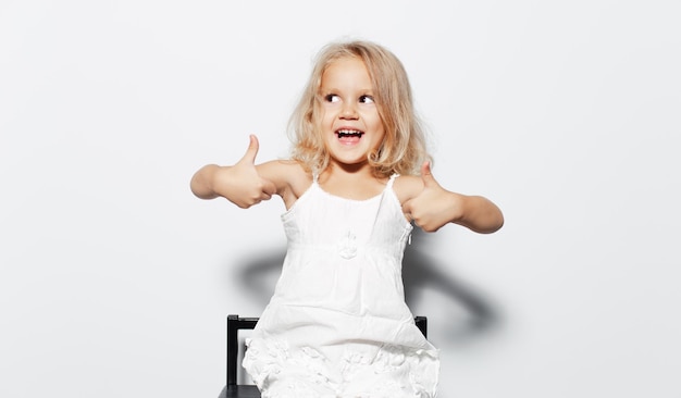 Studio portret van geluk kind meisje met blond haar duimen opdagen op zoek weg op witte achtergrond