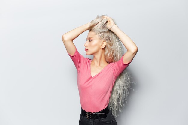 Studio portret van jong blond aantrekkelijk meisje met handen op hoofd in haar pastel roze shirt dragen op witte achtergrond