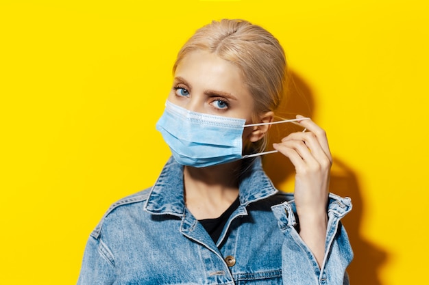 Studio portret van jong blond meisje met blauwe ogen, medische griep masker tegen coronavirus. Achtergrond van gele kleur.