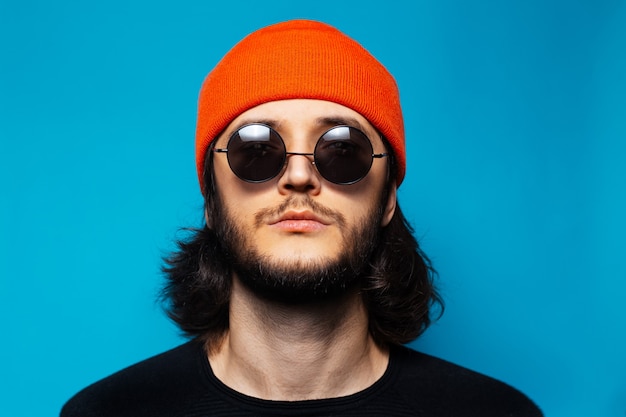 Studio portret van jonge zelfverzekerde man op blauwe achtergrond. man met lang haar met oranje hoed, ronde zonnebril en zwarte trui die omhoog kijkt.