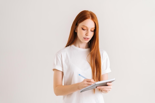 Studio portret van mooie roodharige jonge vrouw schrijven in notitieboekje naar beneden te kijken staande op witte geïsoleerde achtergrond Zelfverzekerde vrouwelijke student maken van aantekeningen in papieren dagboek