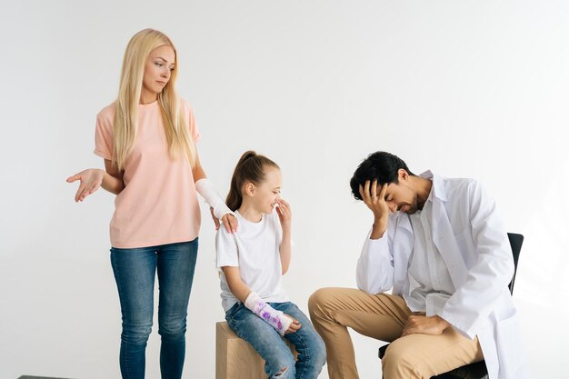 Studio portret van vermoeide mannelijke kinderarts gewonde vrouwelijke patiënt met dochtertje met gebroken hand gewikkeld in gipsverband met grappig gebaar