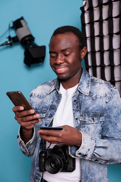 Studio producer making an online payment on smartphone with credit card. Smiling photography enthusiast having DSLR camera making digital purchase with credit card. Studio shot