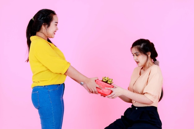 Studio shot of Asian chubby female model in casual wear giving red Birthday Christmas New Year present gift box with gold ribbon to friend or sister who sitting surprising exciting on pink background.