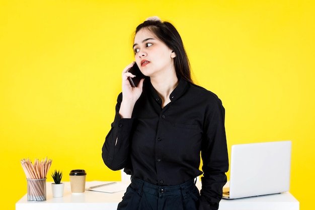 Studio shot of Asian confused amazed doubt thoughtful female businesswoman employee in black outfit sitting on working desk checking news on e-mail from smartphone at office on yellow background.