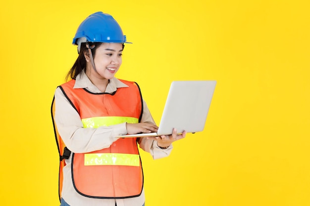 Studio shot of Asian female professional engineering foreman manager wear hard helmet and reflective safety vest stand smiling holding laptop computer working on industrial site on yellow background.