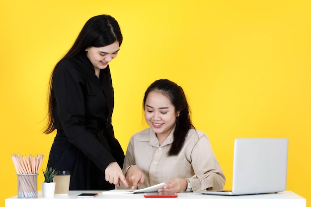 Studio shot of asian happy female mentor employee standing smiling help teaching job training to chubby trainee colleague sitting working with paperwork document at office desk on yellow background