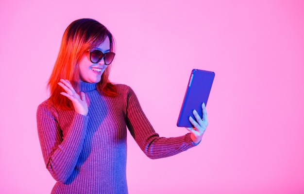 Studio shot of Asian young female model in gray turtleneck sweater black shades standing smiling holding tablet computer video call waving hand say hi hello greeting goodbye on pink light background.