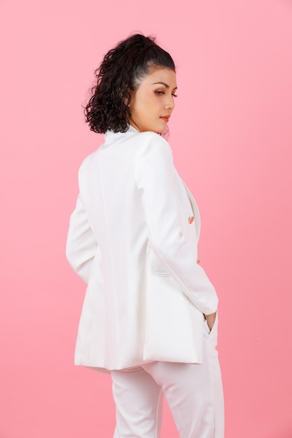 Studio shot of Asian young sexy curly hairstyle professional successful businesswoman in white fashionable casual suit standing posing holding hands in pants pockets turning back on pink background