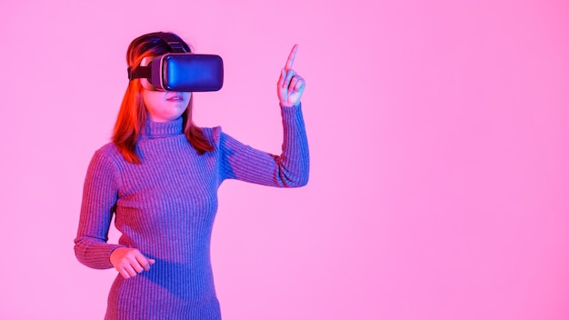 Studio shot of asian young teen female model in gray turtleneck\
dress wears artificial intelligence virtual reality vr video game\
goggles headset playing 3d console gameplay on pink light\
background.
