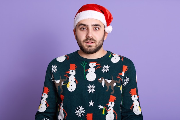 Studio shot of astonished serious man wearing Santa hat and funny pullover with snowflakes standing with opened mouth, looking at camera over blue studio