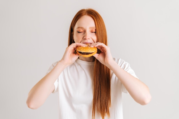 白い孤立した背景に食欲をそそるおいしいハンバーガーのかみ傷を楽しんで目を閉じて魅力的な若い女性のスタジオショット。おいしいハンバーガーを食べる女性のクローズアップ正面図。