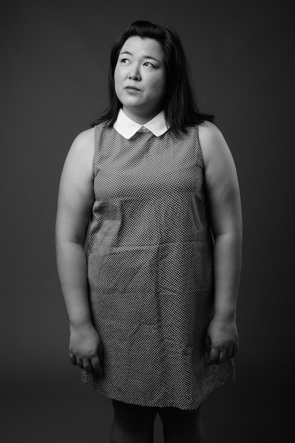 Studio shot of beautiful overweight Asian woman wearing dress against gray background in black and white