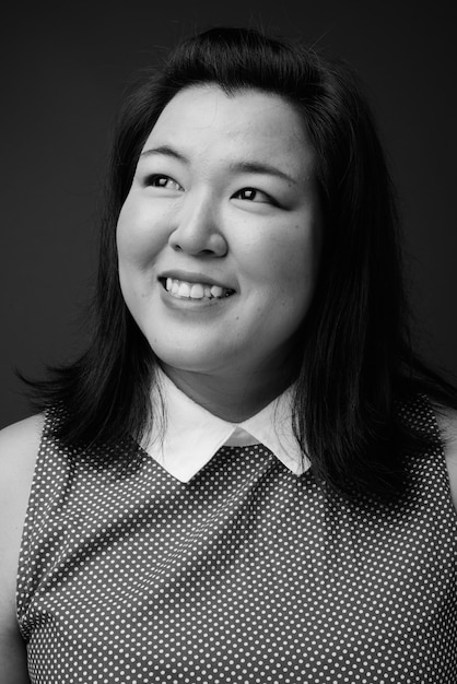 Studio shot of beautiful overweight Asian woman wearing dress against gray background in black and white