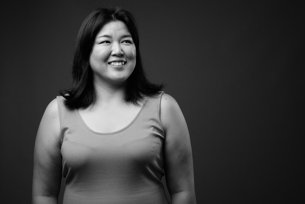 Studio shot of beautiful overweight Asian woman wearing sleeveless dress against gray background in black and white