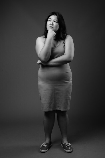 Studio shot of beautiful overweight Asian woman wearing sleeveless dress against gray background in black and white