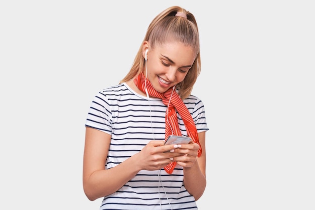 Studio shot of blonde positive smiling young woman using cellphone messaging to her friend Happy female chatting with her boyfriend looking at screen of smart phone listening music in earphones