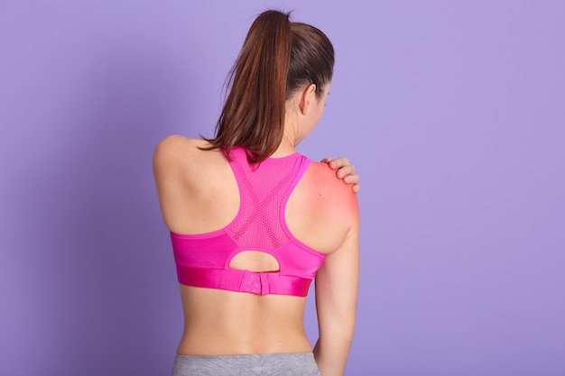 Studio shot of caucasian healthy girl having pain in shoulder, backwards view of sporty female