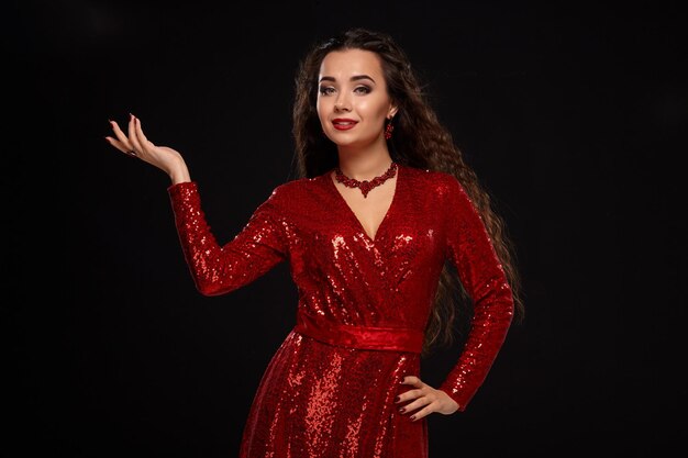 Studio shot of charming young caucasian cute brunette woman in red evening dress posing before camera. Black background.