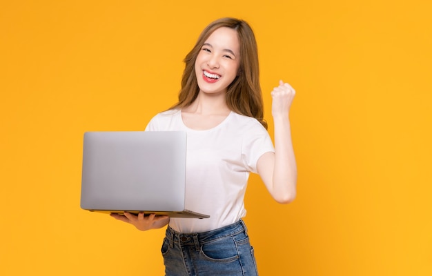 Studio shot of cheerful beautiful Asian woman in white t-shirt and holding laptop with raises arms and fists clenched with shows strong powerful on yellow background.