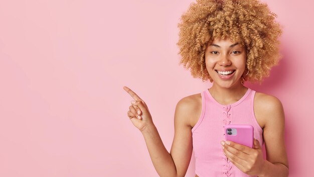 Studio shot of cheerful young european woman with curly hair
holds mobile phone points at blank space for your advertising
content demonstrates place for promotion isolated over pink
background