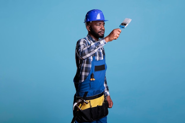 스튜디오 샷 건설 노동자는 집 리모델링을 위해 수리 도구를 사용하여 파란색 배경에 격리된 균일한 보호용 헬멧을 착용합니다. 아프리카 계 미국인 화가 집중.