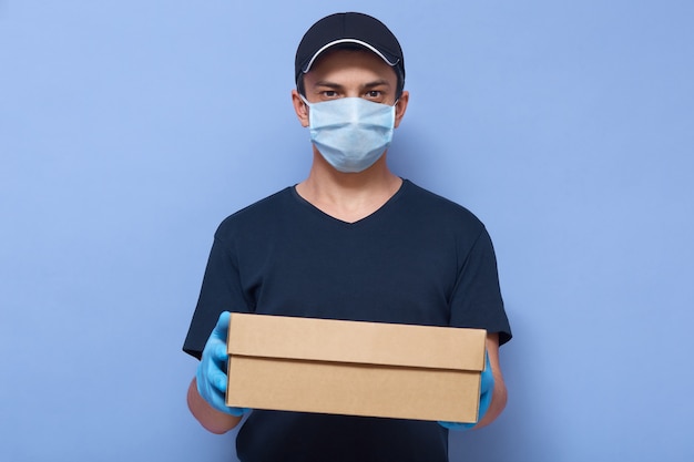 Studio shot of delivery man dresses cap, t shirt, medical mask and latex gloves, wears protective closing while working to protect himself from dangerous corona virus