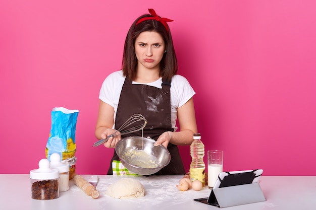 Studio shot of disappointed brunette housewife standing at kitchen with unpleasant facial expression, holding bowl and whisk in both hands, failed to mix all ingredients in proper way, frown face.