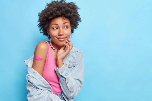 Studio shot of dreamy woman with curly hair looks away shows shoulder with adhesive plaster after getting vaccination wears denim shirt isolated over blue wall