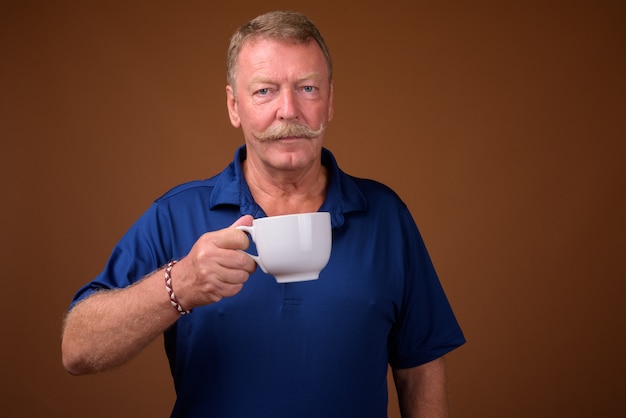 Studio shot of handsome senior man with mustache wearing blue polo shirt against brown