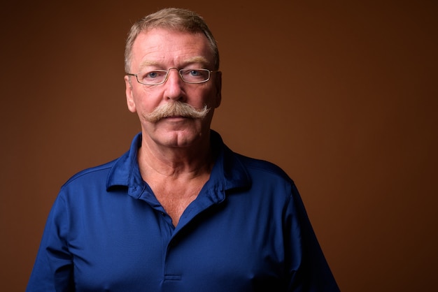 Studio shot of handsome senior man with mustache wearing blue polo shirt and eyeglasses against brown