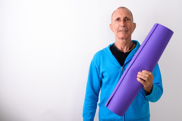 Studio shot of happy bald senior man smiling while holding yoga mat