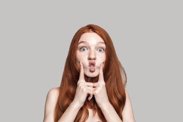 Studio shot of happy young redhead woman making crazy face and grimacing while standing against grey background